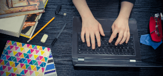 Hands typing on a laptop. Notebooks and textbooks sit beside the laptop.