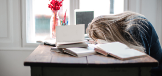 Someone with long blonde hair rests their head on a pile of books. They seem frustrated.